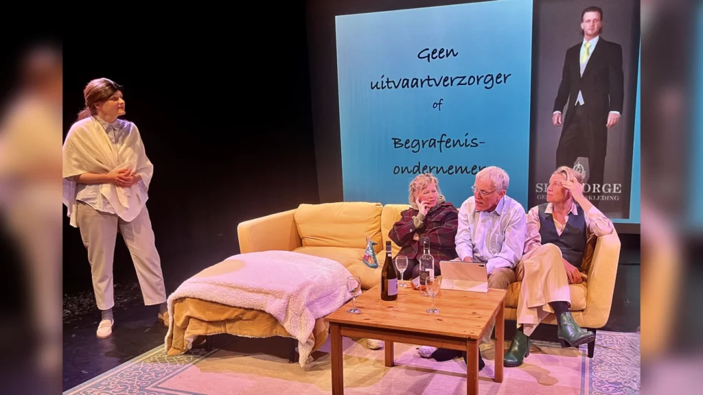 Spelers op een podium met een bank en een scherm met een uitvaartverzorger